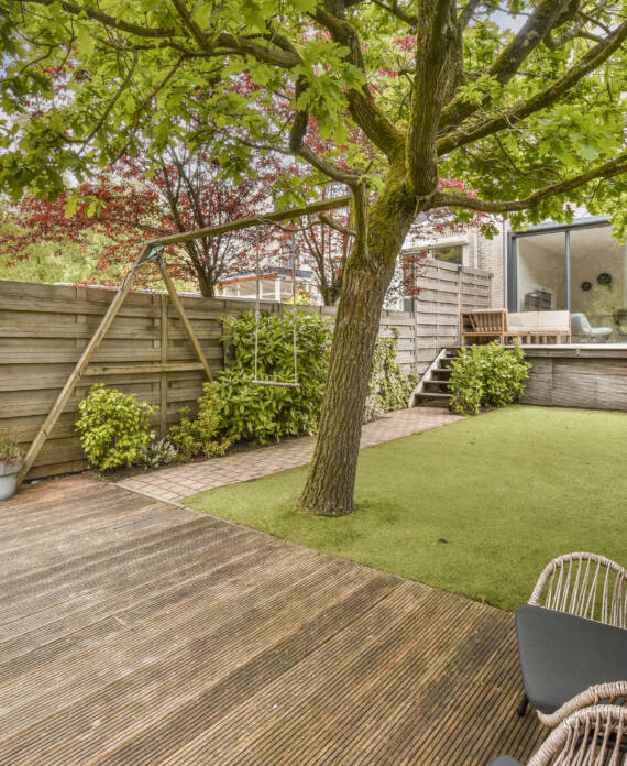 a backyard area with wooden decking and green grass in the yard is surrounded by trees, shrubs and flowers