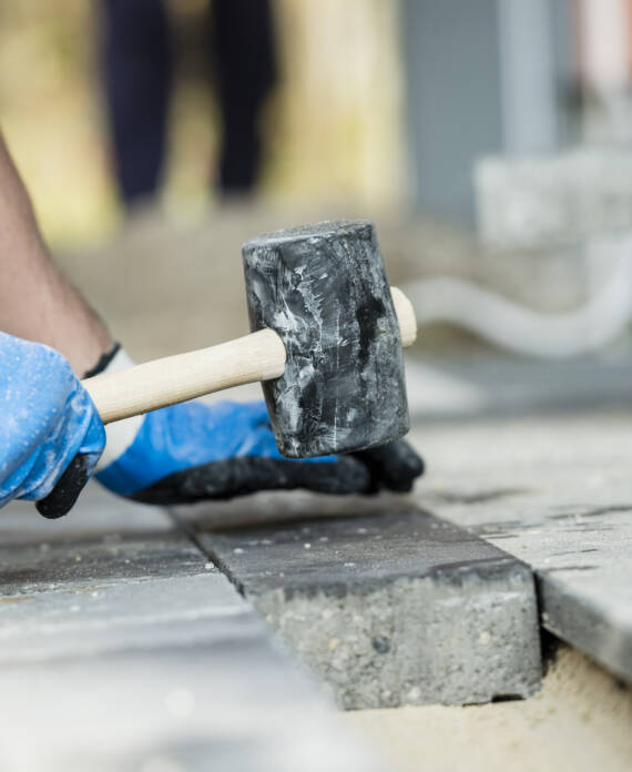 Workman laying a paving stone or brick tamping it down with a large mallet in a close up view of his hands.